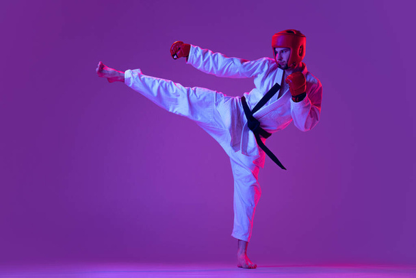 Studio shot of male taekwondo fighter in sports uniform in action isolated over purple background in neon light. Concept of sport, workout, competition, ad - Foto, Bild