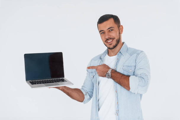 Uses laptop. Young handsome man standing indoors against white background. - Фото, зображення