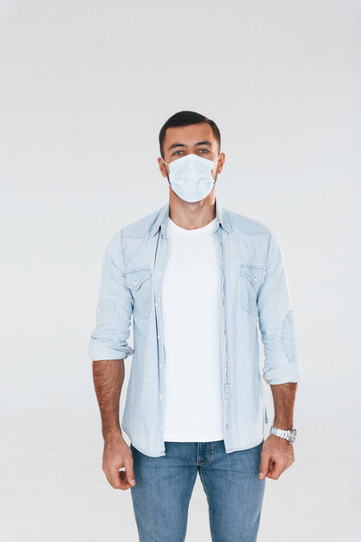 In protective mask. Young handsome man standing indoors against white background. - Valokuva, kuva