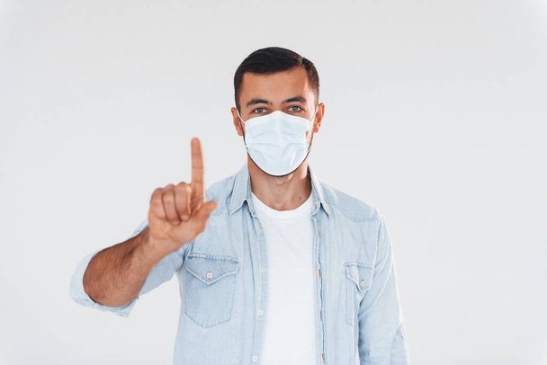In protective mask. Young handsome man standing indoors against white background. - Фото, изображение