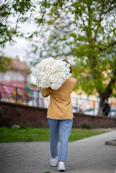 Young brunette female in modern beige outfit with bunch of chryzantem flowers walking in sunny european city street. Back view. Aesthetics. Beautiful girl with big daisies bouquet in field - Фото, зображення