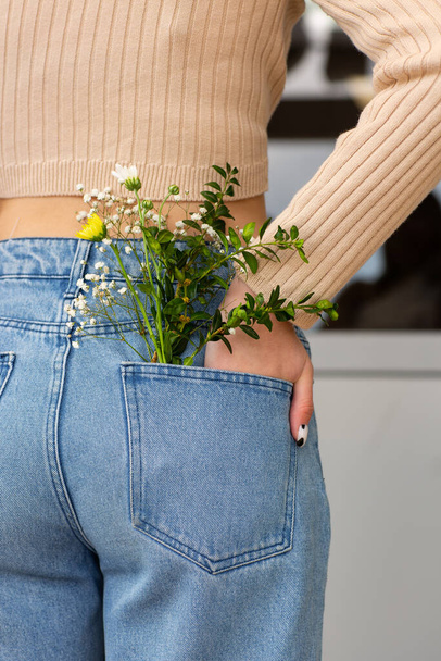Woman with beautiful tender flowers in back pocket of jeans. Woman stabs back to camera. A tiny bouquet of field flowers in a back pocket of women's jeans. - Valokuva, kuva