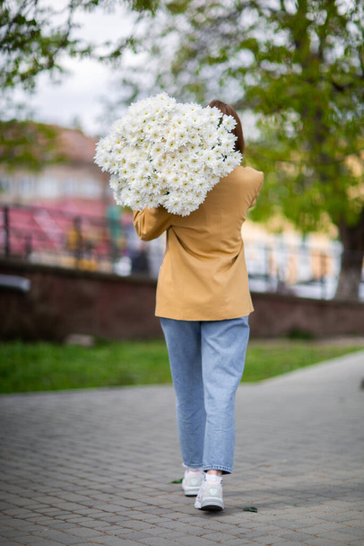 Young brunette female in modern beige outfit with bunch of chryzantem flowers walking in sunny european city street. Back view. Aesthetics. Beautiful girl with big daisies bouquet in field - Foto, Bild