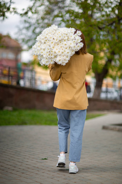 Young brunette female in modern beige outfit with bunch of chryzantem flowers walking in sunny european city street. Back view. Aesthetics. Beautiful girl with big daisies bouquet in field - Valokuva, kuva