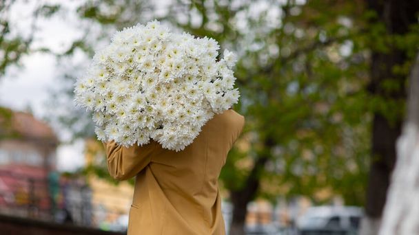 Young brunette female in modern beige outfit with bunch of chryzantem flowers walking in sunny european city street. Back view. Aesthetics. Beautiful girl with big daisies bouquet in field - 写真・画像