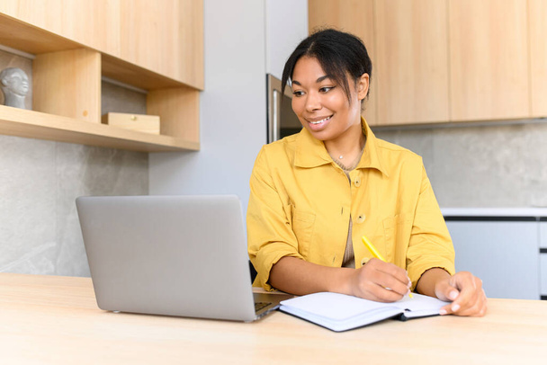 Positive smart young mixed-race female student smiling sitting at the table with laptop in the kitchen, studying from home and taking notes to the notebook, watching an online webinar - Foto, Bild
