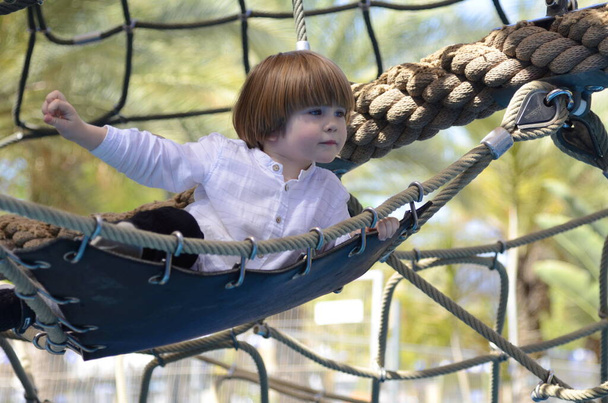Een leuke jongen in een wit shirt ligt in een hangmat. Net op de speelplaats, touwenpark. Web voor activiteiten. Het kind rust uit. - Foto, afbeelding