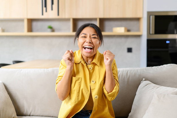 Amazed African-American lady raising hands up yelling yes loud, excited surprised dark skin lady showing win gesture sitting on the sofa at home, looking at the camera - 写真・画像