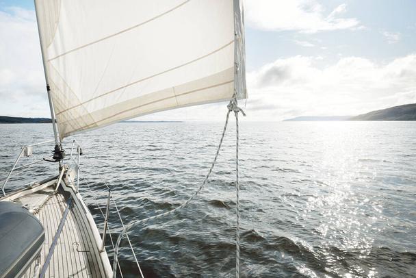 Sloop zmanipuloval moderní jachtu s dřevěnou týkovou palubou plující poblíž skalnatých břehů jezera. Skotsko, Velká Británie. Pohled z paluby na příď, stěžeň, plachty. Wanderlust, volný čas, sport, regata, plavba - Fotografie, Obrázek