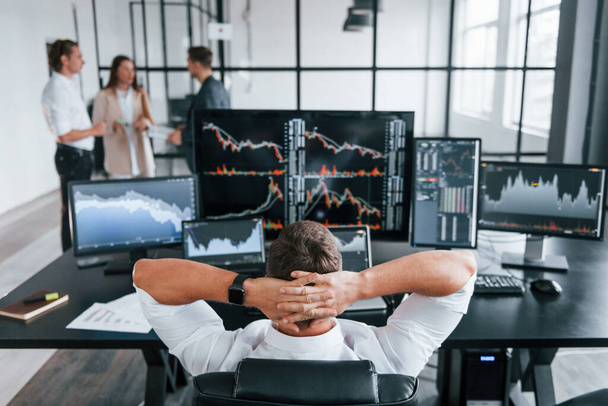 Man sits on chair and takes break. Team of stockbrokers works in modern office with many display screens. - Foto, afbeelding