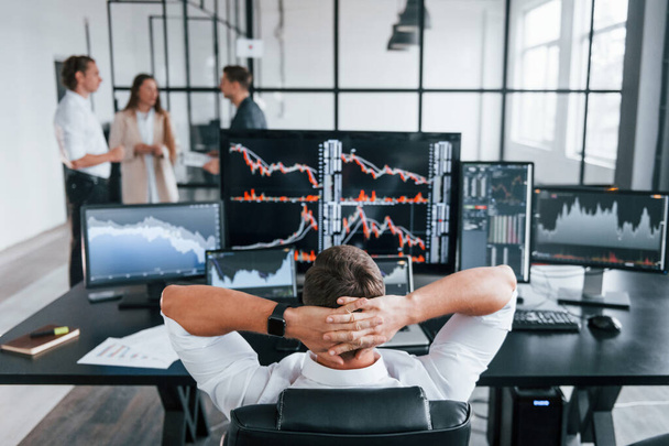 Man sits on chair and takes break. Team of stockbrokers works in modern office with many display screens. - Foto, Imagen