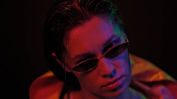 Portrait of young woman wearing jacket and sunglasses in neon color lights . Pretty girl in neon light on black background. Night club, Party concept - 写真・画像