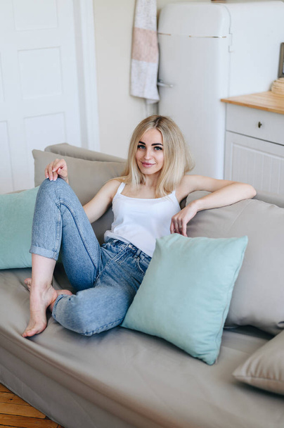 A girl in jeans is sitting on a sofa in the kitchen with her knee bent and looking at the camera. - Foto, Imagen