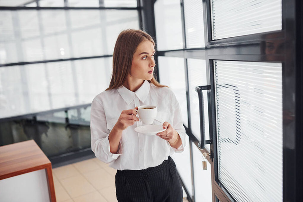 Female office worker in formal clothes standing indoors at daytime with cup of drink. - Foto, afbeelding