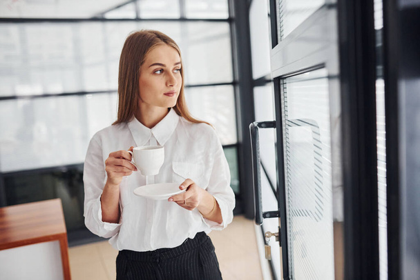 Female office worker in formal clothes standing indoors at daytime with cup of drink. - Foto, Bild