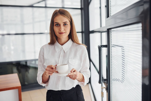 Female office worker in formal clothes standing indoors at daytime with cup of drink. - Фото, зображення
