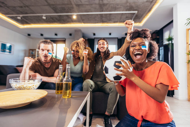 Jonge groep kijken naar sport op televisie en juichen. Emotionele voetbalfans kijken samen naar voetbalwedstrijden thuis. Sport fans met geschilderde gezichten zitten op een bank kijken spel op TV - Foto, afbeelding