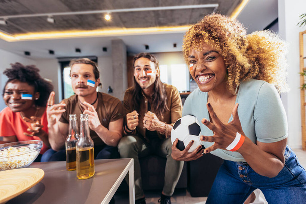 Young group watching sports on television and cheering. Emotional football fans watching football game together at home. Sports Fans with Painted Faces Sitting on a Couch Watch Game on TV - Foto, imagen