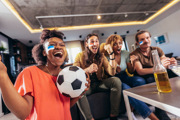 Grupo de jóvenes viendo deportes en la televisión y animando. Fans emocionales del fútbol viendo partidos de fútbol juntos en casa. Abanicos deportivos con caras pintadas sentados en un sofá - Foto, imagen