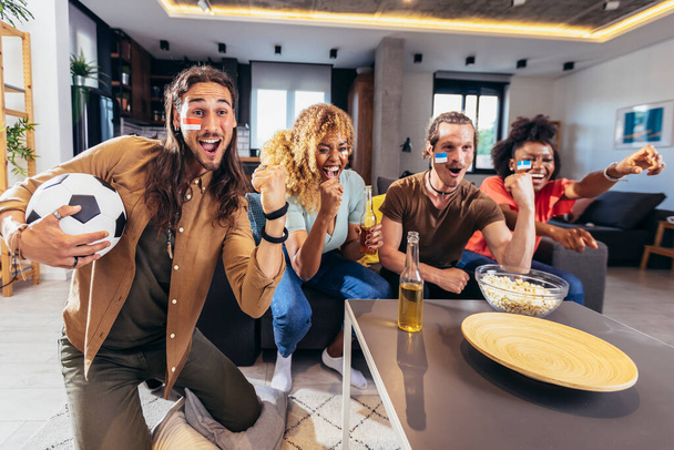 Young group watching sports on television and cheering. Emotional football fans watching football game together at home. Sports Fans with Painted Faces Sitting on a Couch Watch Game on TV - Foto, Imagem