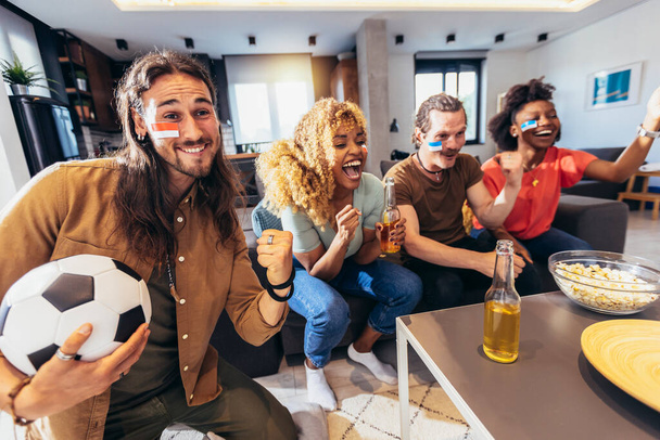 Young group watching sports on television and cheering. Emotional football fans watching football game together at home. Sports Fans with Painted Faces Sitting on a Couch Watch Game on TV - Foto, imagen
