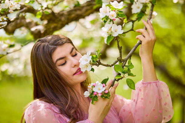 a young woman in a pink dress posing next to a blossoming apple, spring portrait. - Foto, Imagen