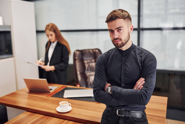 Modern businessman standing indoors in office with his arms crossed. His female colleague behind. - Fotoğraf, Görsel