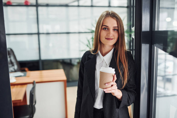 Woman in formal clothes standing indoors in the office with cup of drink. - Foto, Imagen