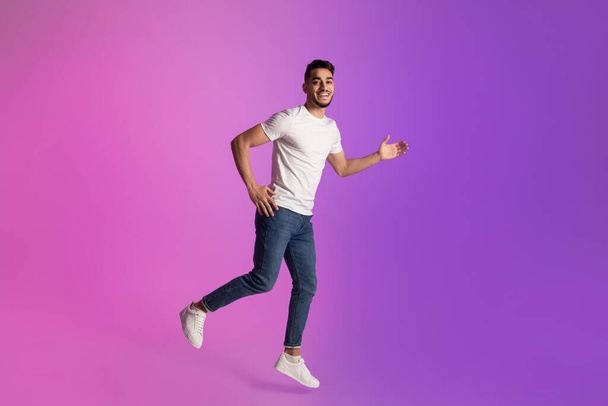 Full length of happy young Arab man in t-shirt and jeans running in neon light, copy space - Zdjęcie, obraz