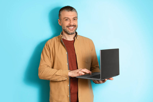 Hombre freelancer de mediana edad que usa el ordenador portátil que trabaja en línea, fondo azul - Foto, Imagen