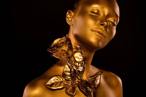 Hot gorgeous lady covered by golden glitter shimmer close her eyes try magical rose flower touch on neck in darkness - Photo, image