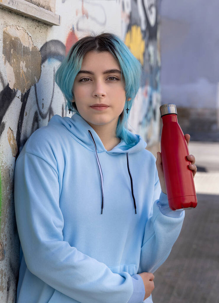 Teenager in blauem Kapuzenpulli mit roter Wasserflasche, die streng aussieht. Blauhaariges Teenie-Mädchen, das sich im Freien in der Nähe von Graffiti-Wänden aufhält. Kleidung und wiederverwendbare Flaschenattrappen. Hipster und Adoleszenz - Foto, Bild