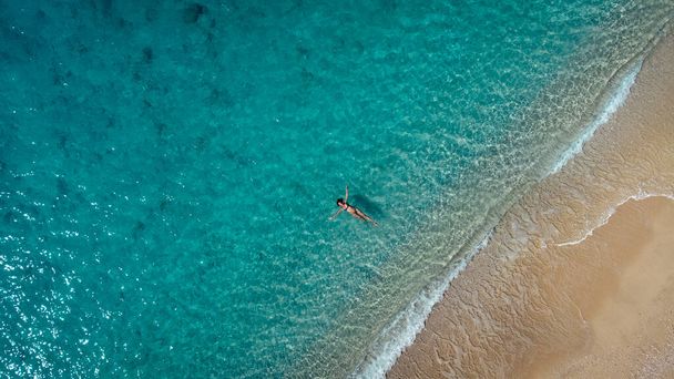 Aerial view of beautiful happy woman in swimsuit laying in the shallow sea water, enjoying sandy beach and soft turquoise ocean wave. Tropical sea in summer season on Egremni beach on Lefkada island. - Zdjęcie, obraz