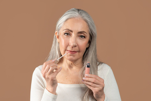 Beauty portrait of senior woman applying lip gloss and looking at camera on brown studio background. Organic makeup - Zdjęcie, obraz