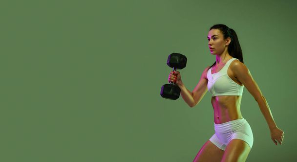 Portrait of young muscular sportive girl training, lifting dumbbell isolated over green studio background in neon light - Fotografie, Obrázek