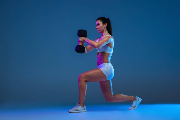 Portrait of yong muscular woman doing squats with heavy dumbbells, training isolated over blue studio background in neon light - Foto, Imagem