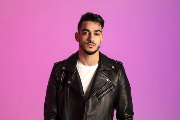 Handsome young Arab guy wearing black leather jacket, posing and looking at camera in neon light. Modern fashion concept - Foto, imagen