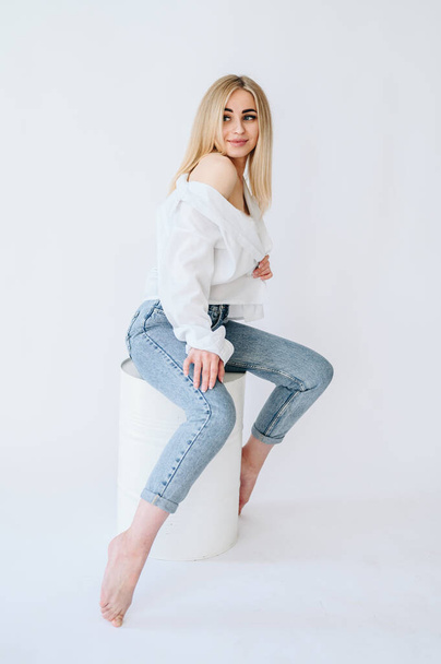 A girl in jeans and a white shirt sits on a white barrel. Blonde girl in jeans on barrel isolated on white background - Fotografie, Obrázek