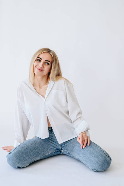 A girl in jeans and a white shirt sits casually on her knees. Blonde woman on a white background in jeans - Φωτογραφία, εικόνα