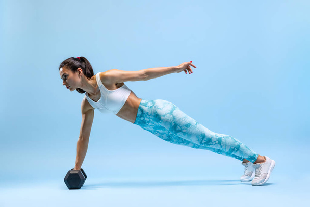 Portrait of young sportive woman standing on plank with dumbbell isolated over blue studio backgtound. Endurance development. - Fotografie, Obrázek