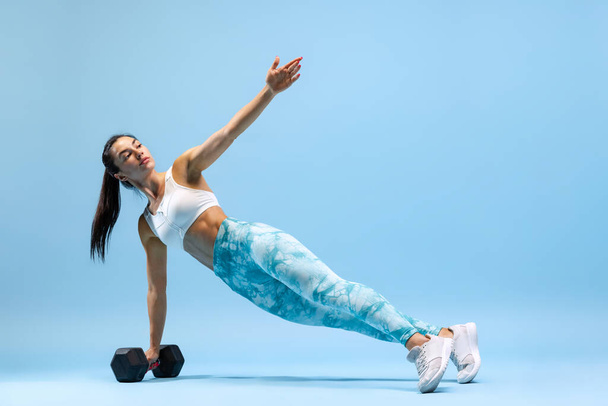 Portrait of young sportive woman training, standing on side plank with dumbbell isolated over blue studio background - Foto, Imagem