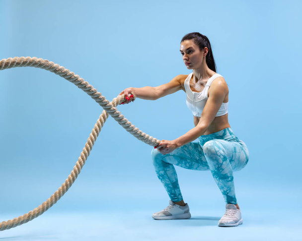 Δυναμικό πορτρέτο της νεαρής αθλητικής γυναίκας κατάρτισης, κάνει ασκήσεις με σχοινί απομονώνονται πάνω από το μπλε φόντο στούντιο - Φωτογραφία, εικόνα
