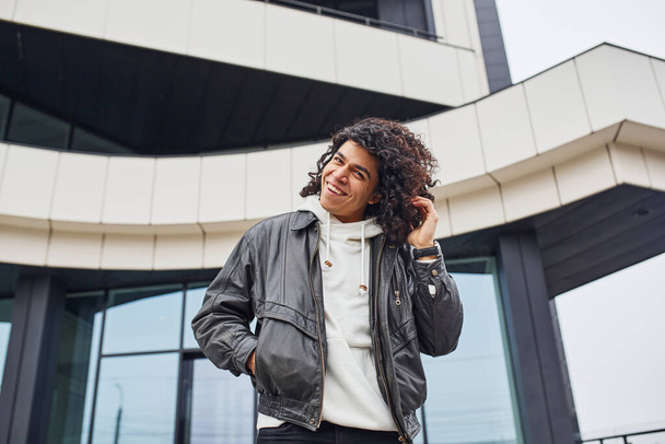 Handsome young man with curly black hair posing for the camera on the street against building. - 写真・画像