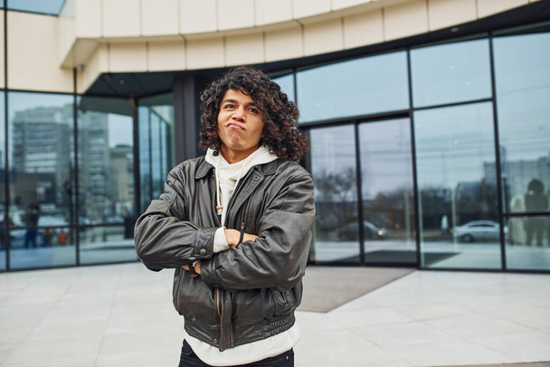 Handsome young man with curly black hair is on the street against building. - Photo, image