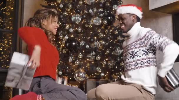 アフリカ系アメリカ人のカップルは、同時にお祭り騒ぎのモミの木の背景にお互いのクリスマスプレゼントを与えます - 映像、動画