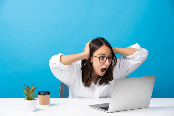 Sorpresa donna ispanica guardando le mani del computer portatile sulla testa mentre seduto dietro la scrivania isolato su sfondo blu. - Foto, immagini