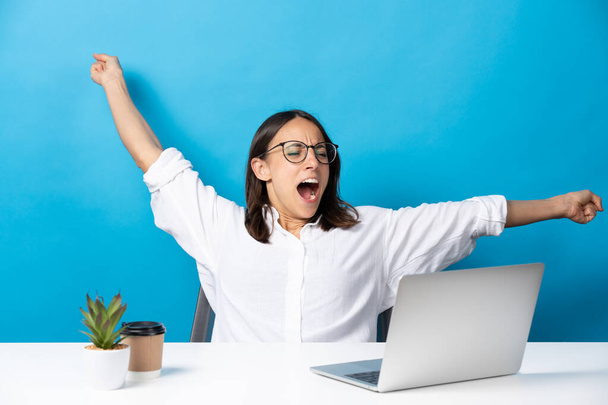 Pretty hispanic businesswoman yawning in front of laptop and sitting behind desk isolated on blue background. - Foto, Imagen