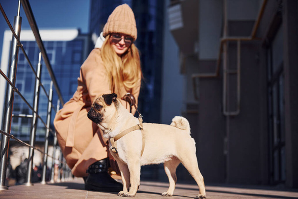 Woman in warm clothes sitting and have fun with her little pug dog near business building that on background. - Zdjęcie, obraz