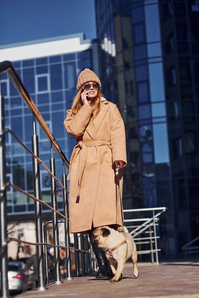 Woman in warm clothes have a walk with her little pug dog near business building that on background and talking by phone. - Φωτογραφία, εικόνα