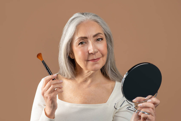Portrét pozitivní starší ženy aplikující přírodní make-up štětcem, při pohledu v miror přes hnědé pozadí studia - Fotografie, Obrázek